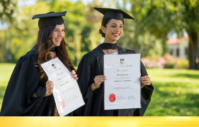 Two international students in graduation robes displaying their diplomas from Adelaide universities